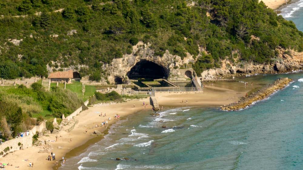 Veduta aerea della Grotta di Tiberio a Sperlonga, con un'ampia insenatura naturale nella roccia ai piedi di una collina verde, vasche archeologiche romane all'ingresso della caverna, la spiaggetta dorata e le acque trasparenti del mare