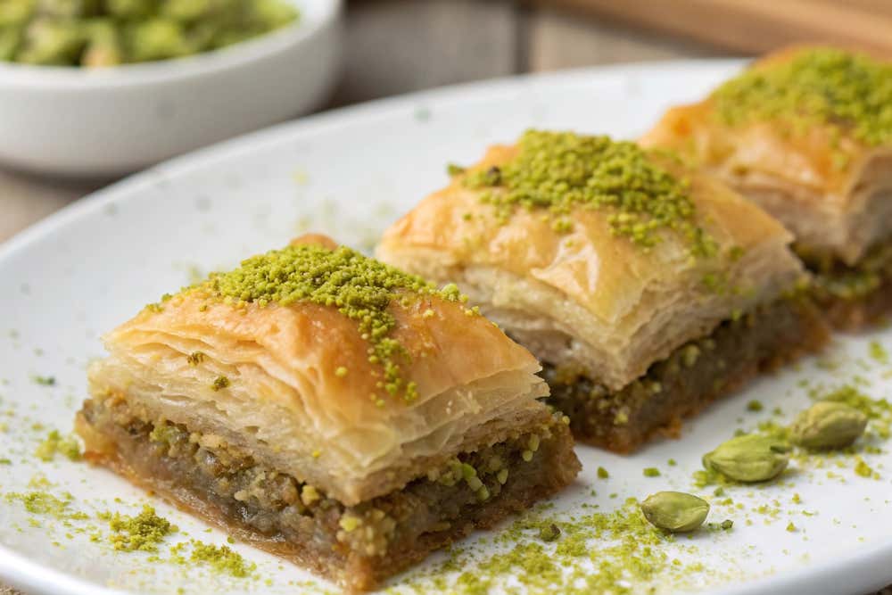 Tre porzioni quadrate di baklava disposte in fila su un piatto bianco, composte da strati sottili di pasta fillo dorata e mista a sciroppo, ripene di frutta secca e guarnite in superficie con granella di pistacchio verde