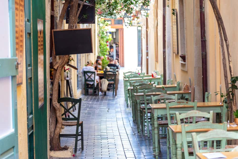 Tavolini e sedie di legno verde allineati all'aperto lungo un vicolo pedonale greco, con persone sedute a conversare rilassate in fondo alla via sotto l'ombra degli alberi