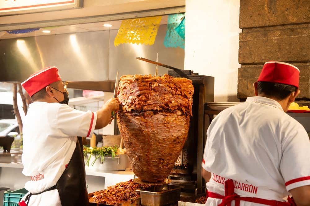 A man wearing a red hat and a black face mask slicing meet from a spit of al Pastor meat.