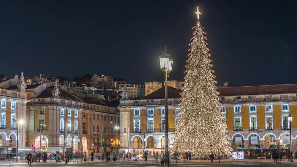 Praça do Comércio, em Lisboa, decorada para o Natal, com uma grande árvore, luzes festivas e a movimentação de pessoas à noite, em meio à arquitetura histórica da cidade