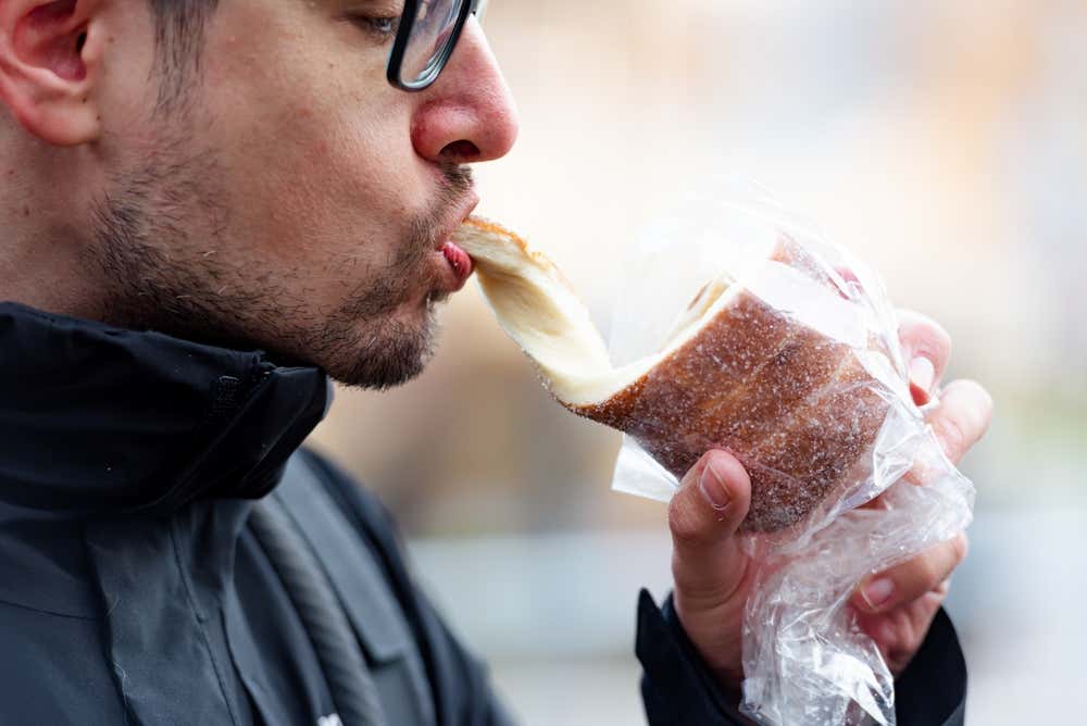 Primo piano di un uomo con occhiali che morde un kürtőskalács (torta camino) caldo, mostrando il morbido impasto interno che si allunga mentre lo stacca dal dolce cilindrico ricoperto di zucchero e cannella