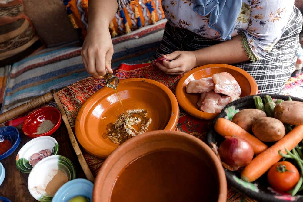 Primer plano desde arriba de una mujer vertiendo aceite de oliva sobre las especias dentro de la base de un tajín de barro, rodeada de ingredientes para la preparación del plato, entre ellos trozos de pollo crudo, cebollas, patatas, zanahorias, tomates y cuencos con varias especias