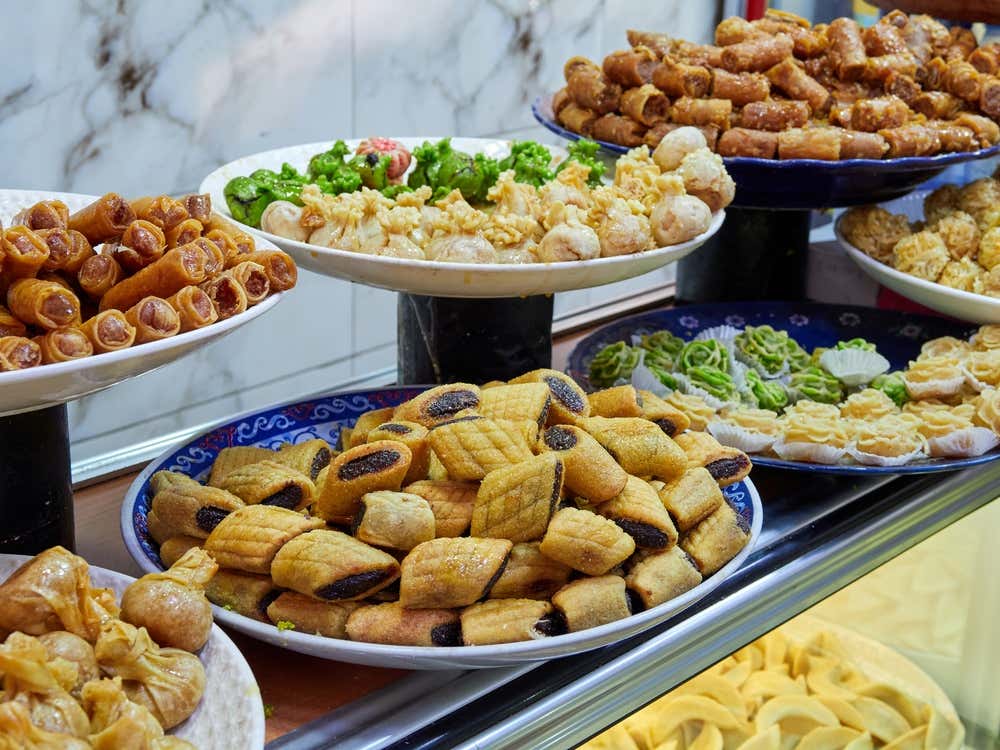 Un amplio escaparate de repostería árabe y marroquí con varios platos de dulces tradicionales de miel, almendra y dátil, entre ellos makroud en forma de rombo en el centro, rollitos (briouates), fardos y dulces decorados con pistacho
