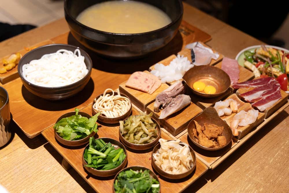 Vista dall'alto degli ingredienti per i noodles Guoqiao mixian, con un grande recipiente di brodo bollente, noodles di riso bianchi e numerosi piattini in legno e rame con carne, gamberi, uova e verdure fresche disposti su un vassoio
