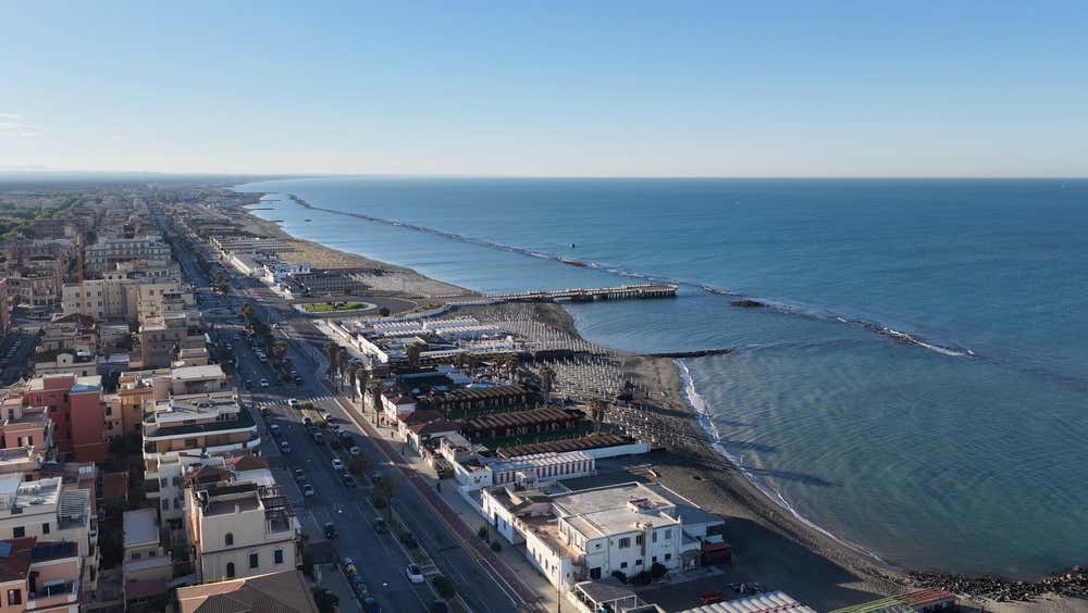 Veduta aerea panoramica del lungomare e della costa di Ostia Lido, con gli stabilimenti balneari, il pontile che si protende nel mar Tirreno blu, la strada costiera affiancata dagli edifici della città e il rettilineo delle spiagge sotto un cielo limpido