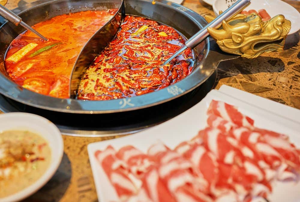 Vista dall'alto di un pentolone diviso a metà (Hot Pot cinese) decorato con testa di drago dorata, contenente due brodi rossi piccanti affioranti di peperoncini, affiancato da un piatto di sottili fette di carne cruda arrotolata