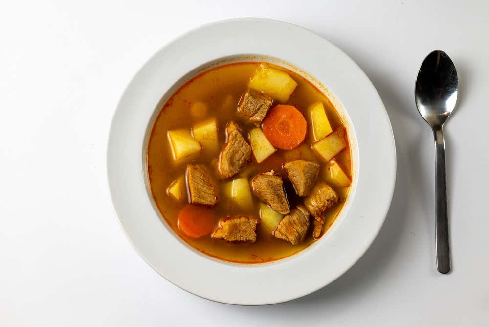 **Alt Text (Testo Alternativo)**

> **Alt text:** Vista dall'alto di un piatto fondo bianco contenente uno stufato denso o zuppa di carne a bocconcini con patate e carote a rondelle nel suo brodo, affiancato da un cucchiaio di metallo su fondo bianco.