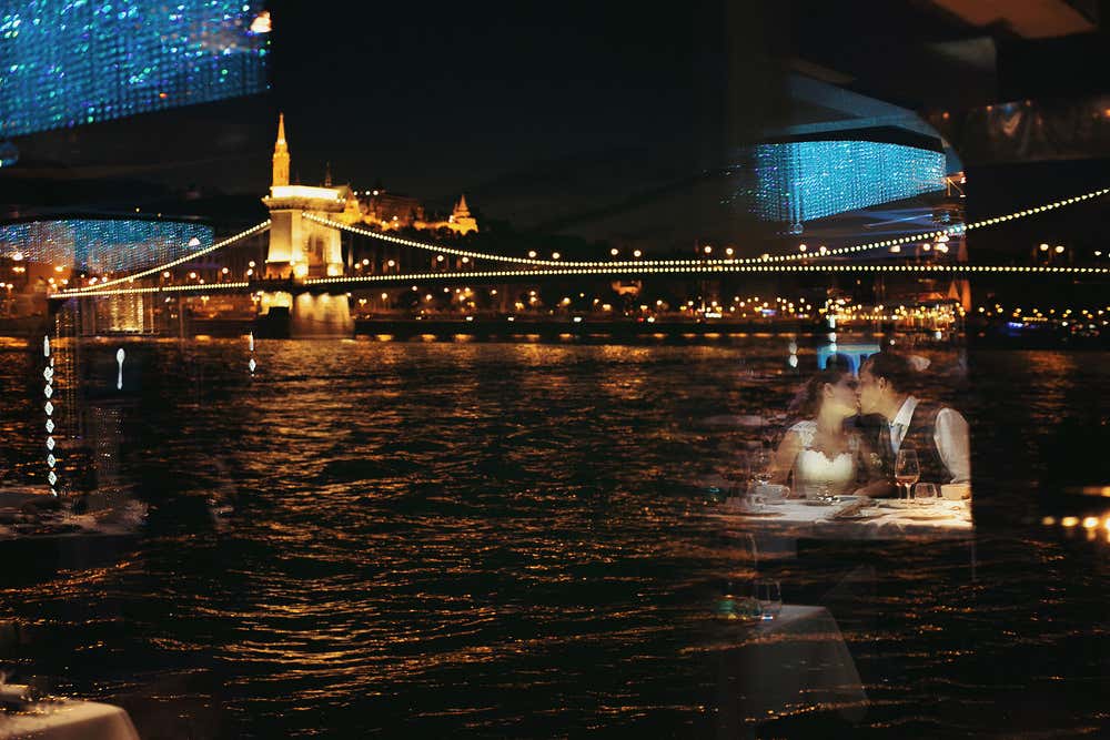 Vista notturna del Ponte delle Catene illuminato a Budapest riflesso sulle acque del Danubio, con il riflesso del vetro in primo piano che mostra una coppia di sposi che si bacia durante una cena romantica