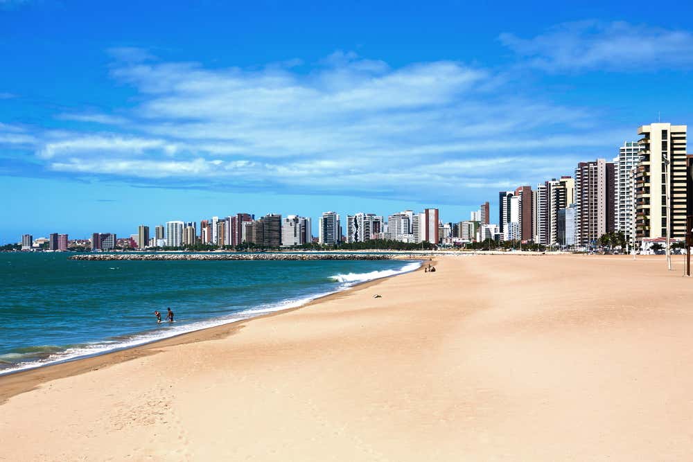Vista da praia de Fortaleza, no Ceará, Brasil, com faixa de areia dourada, mar azul e a orla urbana marcada por altos edifícios que compõem a paisagem da cidade