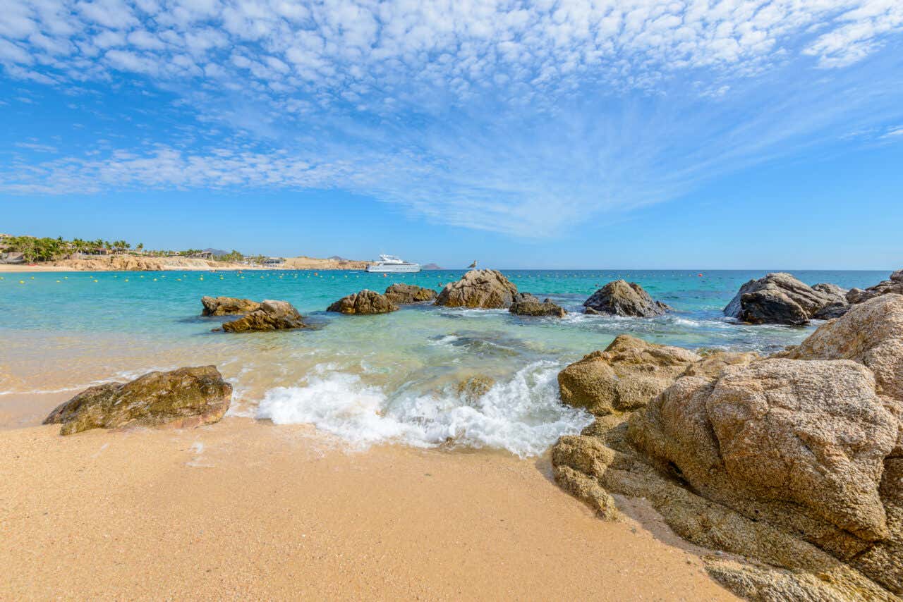 A beach in Los Cabos, the sea is splashing over the rocks and a cruise ship is visible in the background.