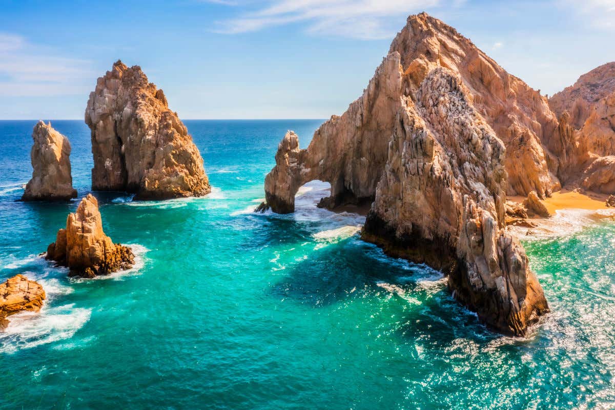 Vista panorámica desde el agua de "El Arco", la famosa formación rocosa natural en Fin de la Tierra, Los Cabos. Las towering rocas son de color beige, y las olas del océano turquesa y azul profundo chocan suavemente contra ellas. El cielo es azul con nubes ligeras
