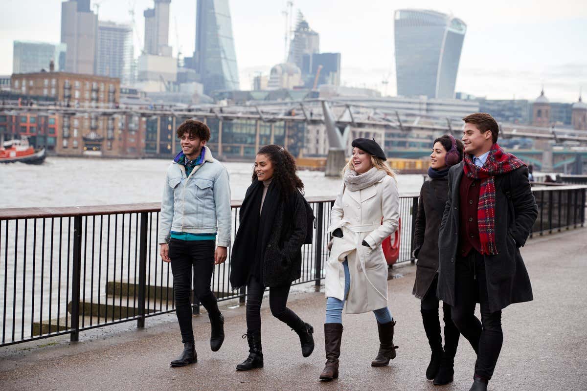 Grupo de amigos bem agasalhados passeando à beira do rio Tâmisa, com o horizonte de Londres ao fundo