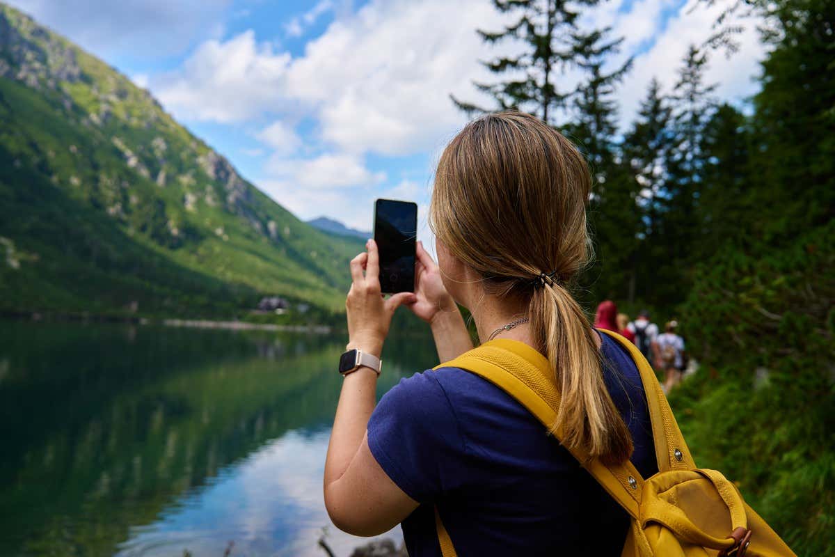 Una senderista con coleta fotografía con el móvil un lago tranquilo rodeado de montañas y pinos