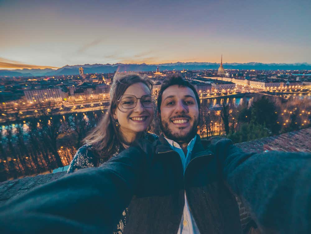 Una coppia sorridente si scatta un selfie al tramonto dal Monte dei Cappuccini, a Torino. Alle loro spalle si vede la città illuminata, con la Mole Antonelliana, il fiume Po e l'arco delle Alpi all'orizzonte, sotto un cielo sfumato tra l'arancio e il viola