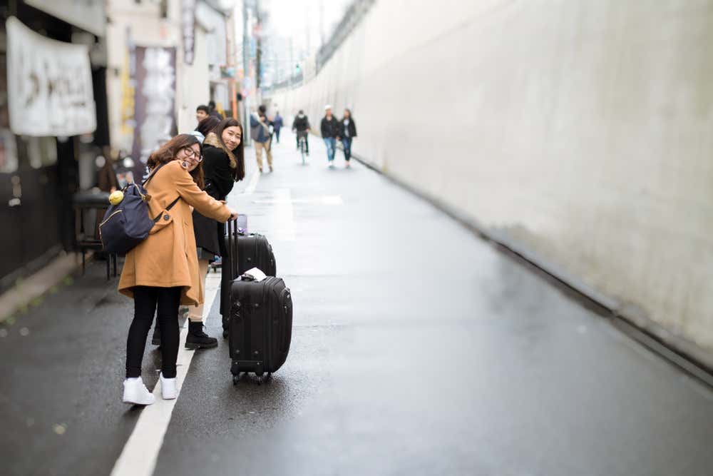 Due ragazze sorridenti con zaino e trolley si voltano verso l'obiettivo in una stretta via di una città giapponese. L'asfalto è bagnato di pioggia e lungo la strada ci sono insegne di negozi. Sullo sfondo, sfocati, passano altri pedoni e un ciclista