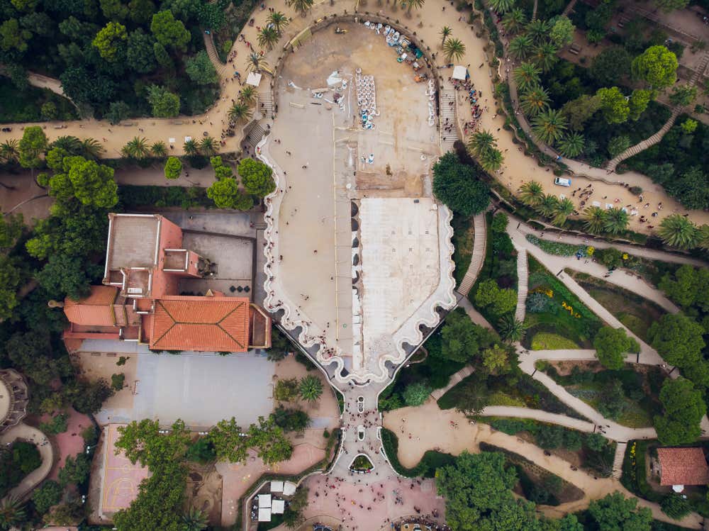 Vista aerea del Parc Güell a Barcellona. Al centro c'è la Plaça de la Natura, con il bordo ondulato della panchina in trencadís, circondata da palme, alberi, vialetti sinuosi e aiuole. Sulla sinistra si vede un edificio dal tetto in tegole arancioni