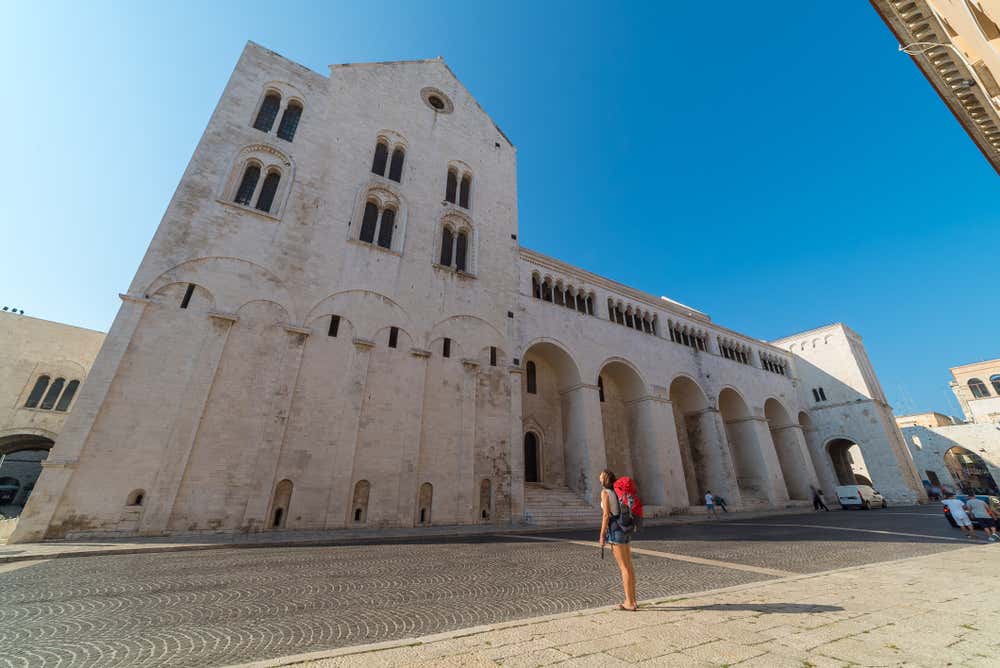 Una turista con uno zaino rosso osserva l'imponente facciata in pietra bianca della Basilica di San Nicola a Bari Vecchia sotto un cielo azzurro e limpido