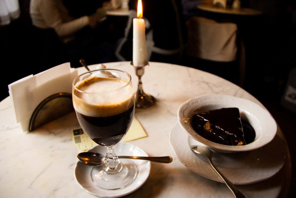 Un vaso de bicerin turinés con capas de espresso, chocolate caliente y crema de leche, servido en la mesa de mármol de un café histórico junto a una vela encendida