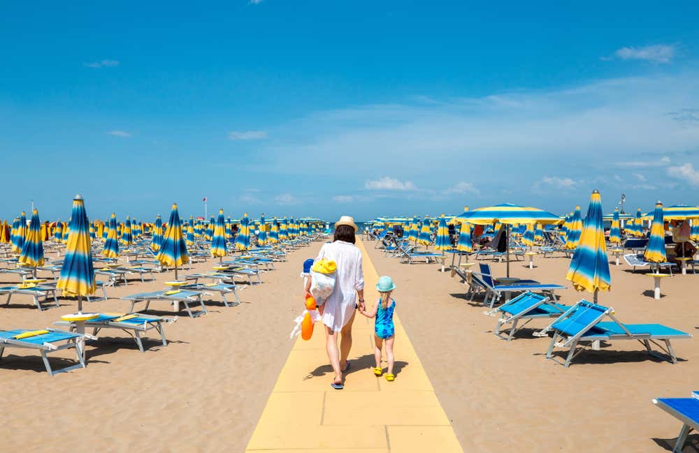 Una madre e la sua bambina camminano per mano lungo una passerella gialla sulla spiaggia di Rimini, circondate da file di ombrelloni e lettini azzurri e gialli sotto un cielo sereno