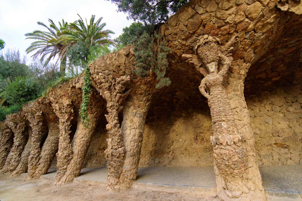 Il Portico della Lavandaia al Parc Güell di Barcellona: una galleria in pietra grezza con colonne inclinate che ricordano tronchi d'albero. In primo piano, una delle colonne è scolpita a forma di donna che porta un cesto di panni sulla testa. Sopra il portico crescono edera, palme e vegetazione mediterranea