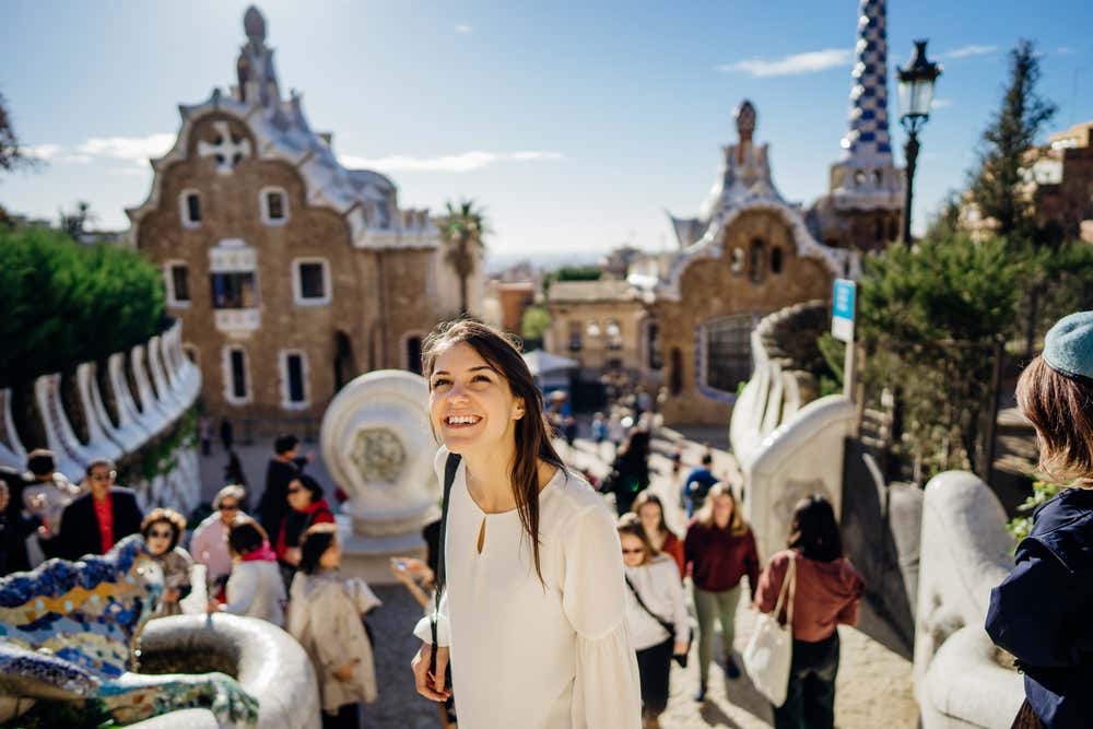 Una giovane turista sorridente sulla Scalinata del Drago del Parc Güell a Barcellona, che guarda verso l'alto in una giornata di sole. Dietro di lei ci sono altri visitatori e i padiglioni d'ingresso di Gaudí, con i tetti in trencadís e la torre a scacchi bianchi e blu. In basso a sinistra si intravede il mosaico colorato del drago