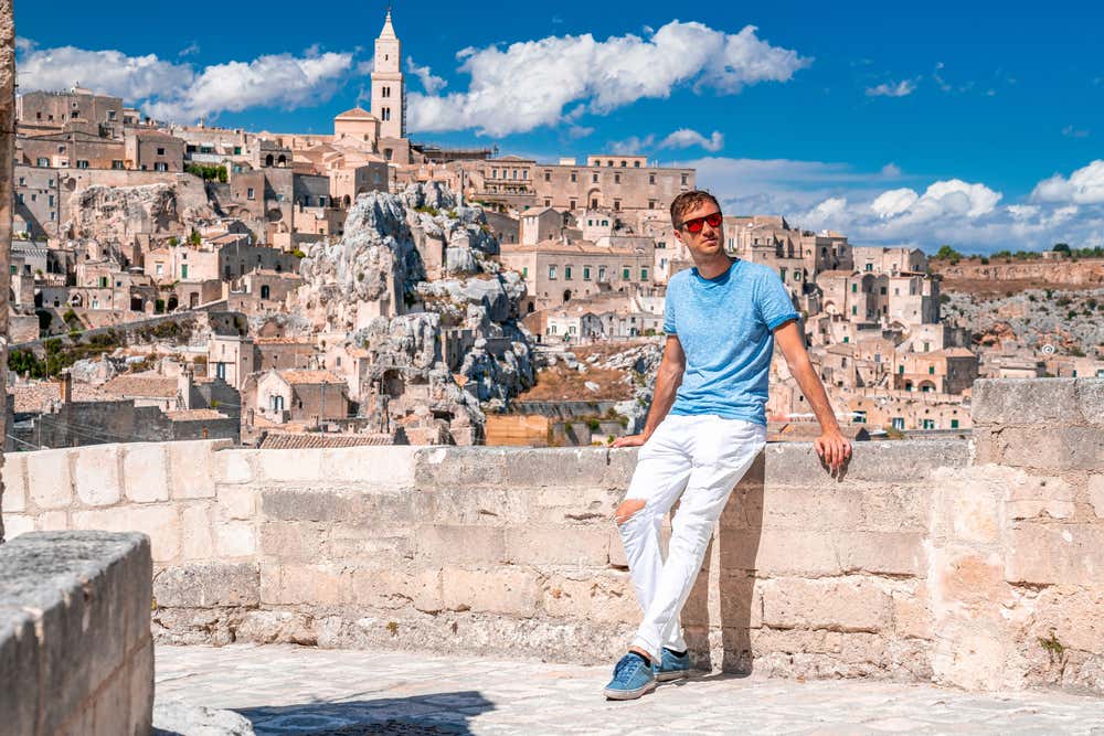 Un turista con maglietta azzurra e occhiali da sole appoggiato a un muretto in pietra, con lo sfondo panoramico dei Sassi di Matera e del campanile della Cattedrale sotto un cielo sereno