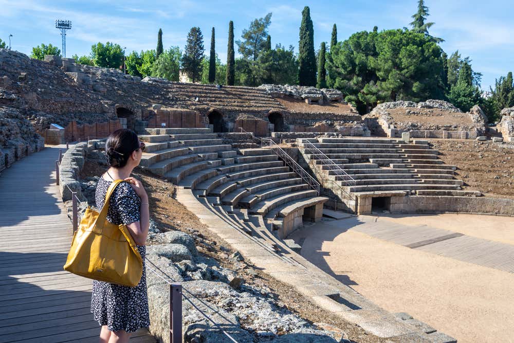 Una donna di profilo con vestito a fiori e borsa gialla osserva le gradinate in pietra dell'antico anfiteatro romano di Mérida, circondato da alberi e cipressi sotto un cielo limpido