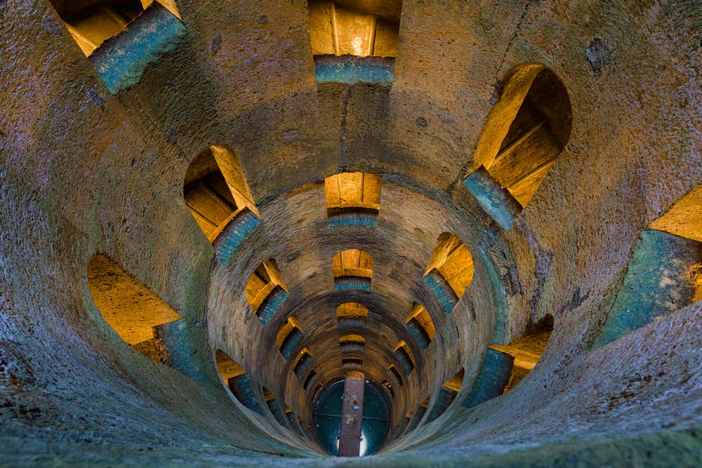 L'interno del Pozzo di San Patrizio a Orvieto fotografato dall'alto verso il fondo. Le pareti cilindriche in pietra sono attraversate da finestre ad arco disposte a spirale, illuminate da una luce calda. In fondo al pozzo si vedono l'acqua e un piccolo ponte