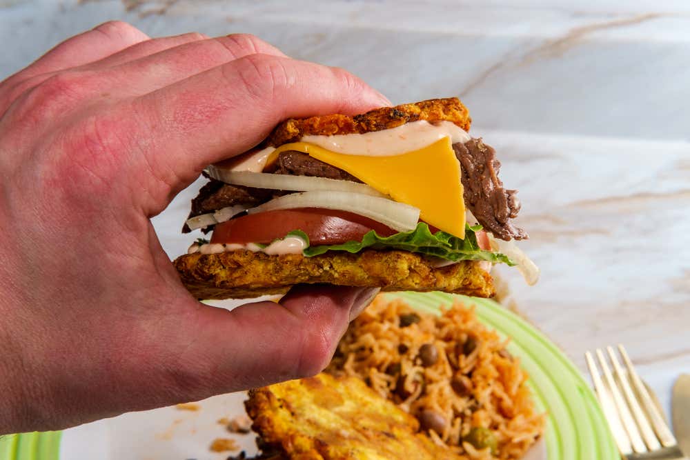 Someone holding a jibarito with cheese, sauce, onion, tomato, lettuce and beef.