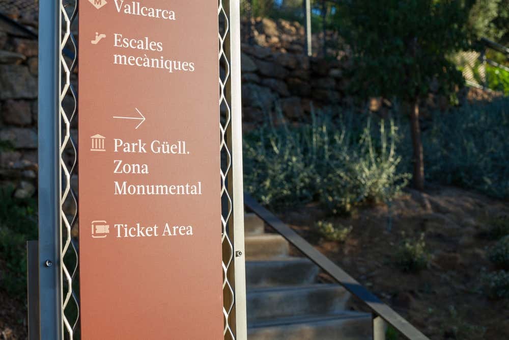 Un cartello color terracotta nel Parc Güell di Barcellona indica la metro Vallcarca, le scale mobili ("Escales mecàniques"), la Zona Monumentale e la biglietteria ("Ticket Area"). Accanto al cartello ci sono una scalinata in pietra con il corrimano e un muretto a secco circondato da vegetazione