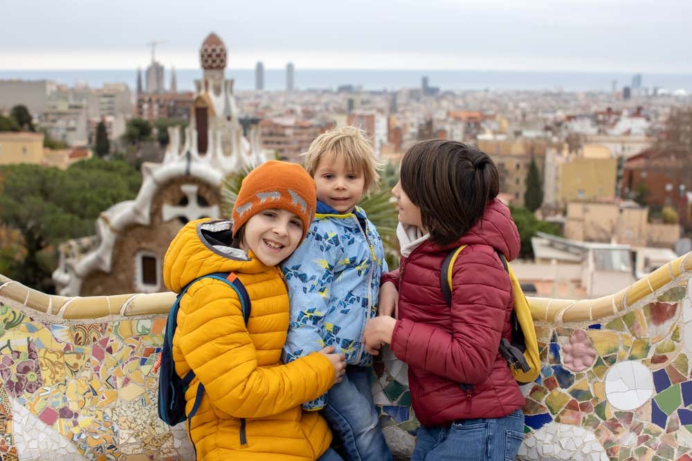 Tre bambini con giacche colorate e zainetti sorridono davanti alla panchina ondulata in trencadís della Plaça de la Natura, al Parc Güell di Barcellona. Alle loro spalle si vedono uno dei padiglioni d'ingresso di Gaudí e il panorama della città fino al mare, in una giornata nuvolosa