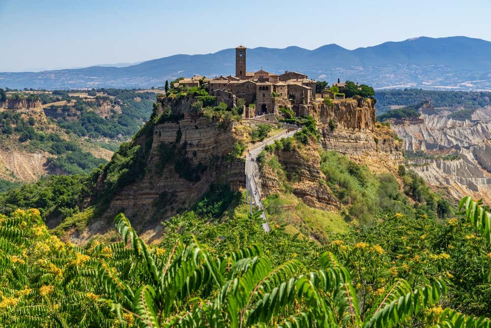 Civita di Bagnoregio arroccata su uno sperone di tufo, con le case in pietra e il campanile che svettano sulla cima. Un lungo ponte pedonale in salita collega il borgo al resto del paesaggio. Tutto intorno si stendono i calanchi, colline d'argilla erose, e sullo sfondo ci sono montagne sotto un cielo azzurro. In primo piano si vedono fronde verdi