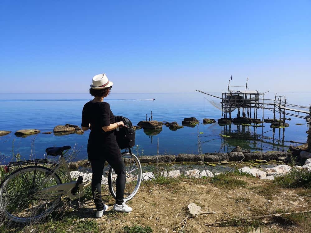 Una donna con un cappello di paglia e una bicicletta guarda il mare calmo e azzurro della Costa dei Trabocchi, in Abruzzo. Sulla destra si vede un trabocco, l'antica macchina da pesca in legno costruita su palafitte, con scogli affioranti e cielo limpido