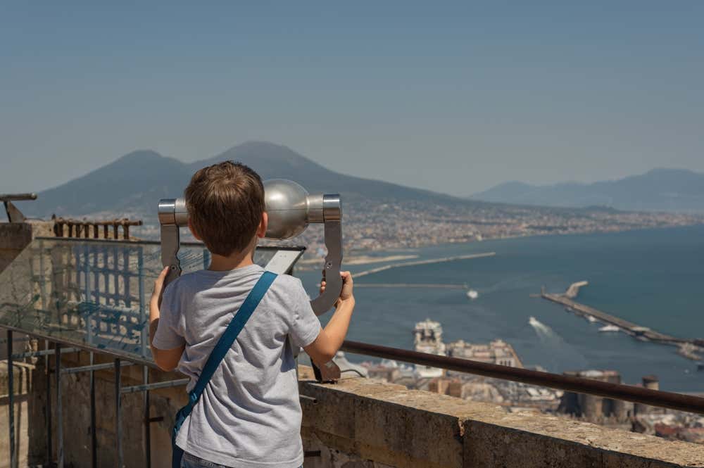 Un bambino visto di spalle guarda attraverso un binocolo panoramico il Golfo di Napoli e il Vesuvio da una terrazza panoramica della città