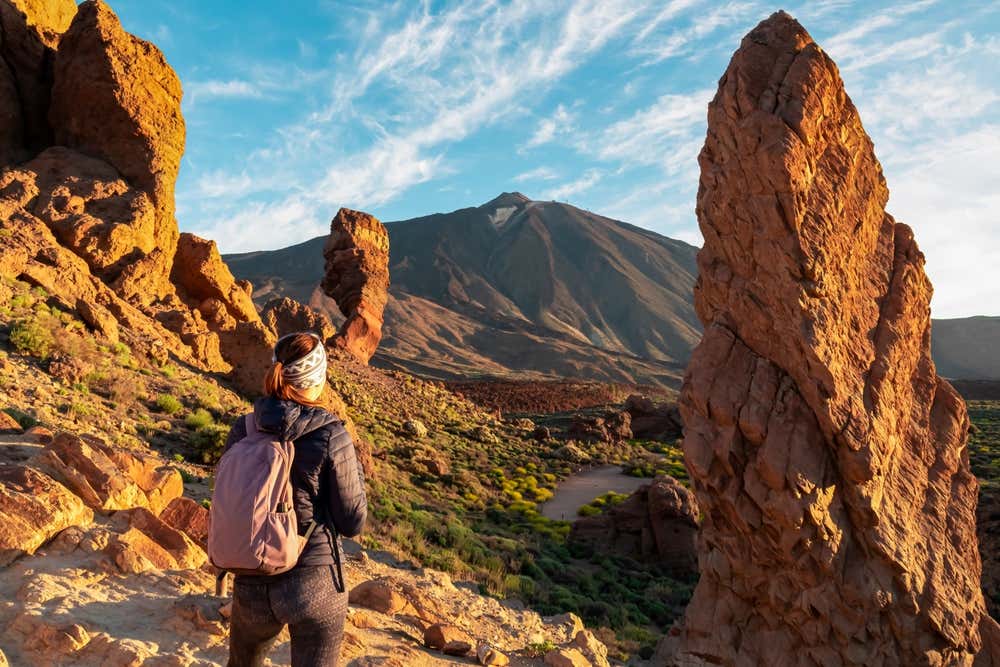 Un'escursionista di spalle con uno zaino rosa ammira le imponenti formazioni rocciose vulcaniche dei Roques de García e il picco del Teide sullo sfondo nel Parco Nazionale del Teide a Tenerife