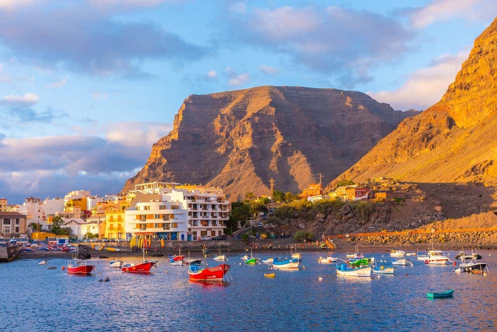 Vista dal mare del porto e del villaggio di Valle Gran Rey a La Gomera, con barche da pesca colorate ancorate nell'acqua azzurra ed imponenti scogliere rocciose illuminate dalla luce calda del tramonto
