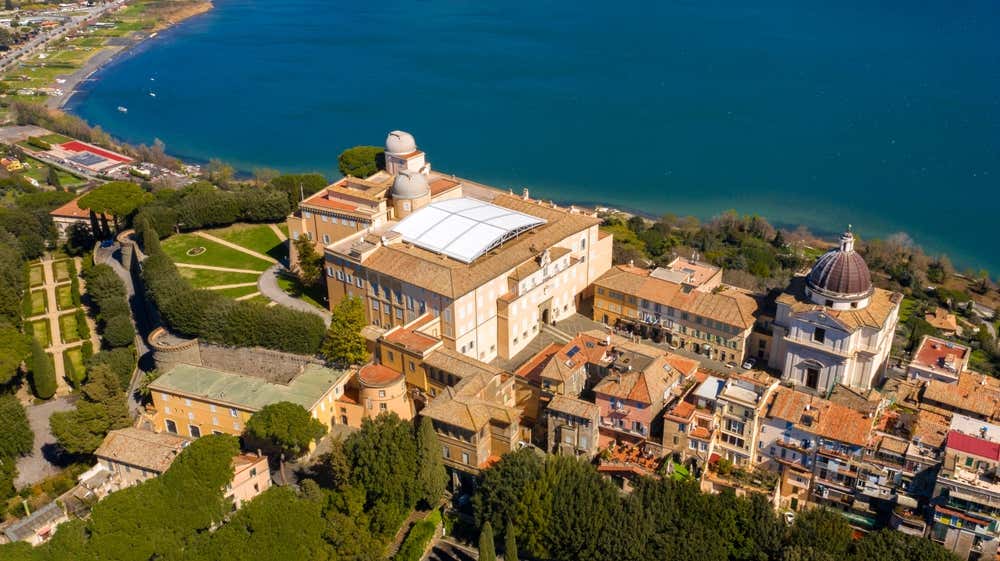 Vista aerea di Castel Gandolfo, sui Colli Albani. Al centro c'è il Palazzo apostolico con le cupole dell'antica Specola vaticana, sulla destra la chiesa con la cupola di Bernini e intorno le case del borgo e i giardini. Sullo sfondo si apre il lago di Albano, di un blu intenso