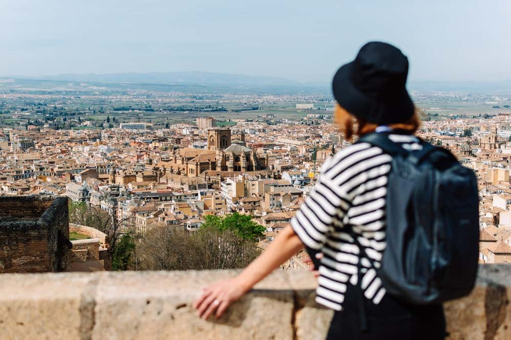Una turista di spalle con cappello nero, maglia a righe e zaino scuro guarda da un belvedere in pietra il panorama dall'alto della città di Granada, con i suoi tetti storici e la cattedrale in evidenza