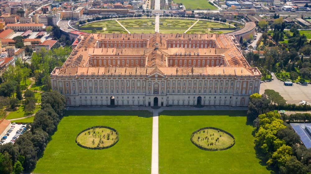 Vista aerea della Reggia di Caserta, l'imponente palazzo reale dei Borbone. Dall'alto si vedono la lunga facciata e i quattro cortili interni. In primo piano ci sono i prati del parco con due aiuole circolari, mentre sullo sfondo si apre la piazza ellittica con i giardini geometrici e la città intorno