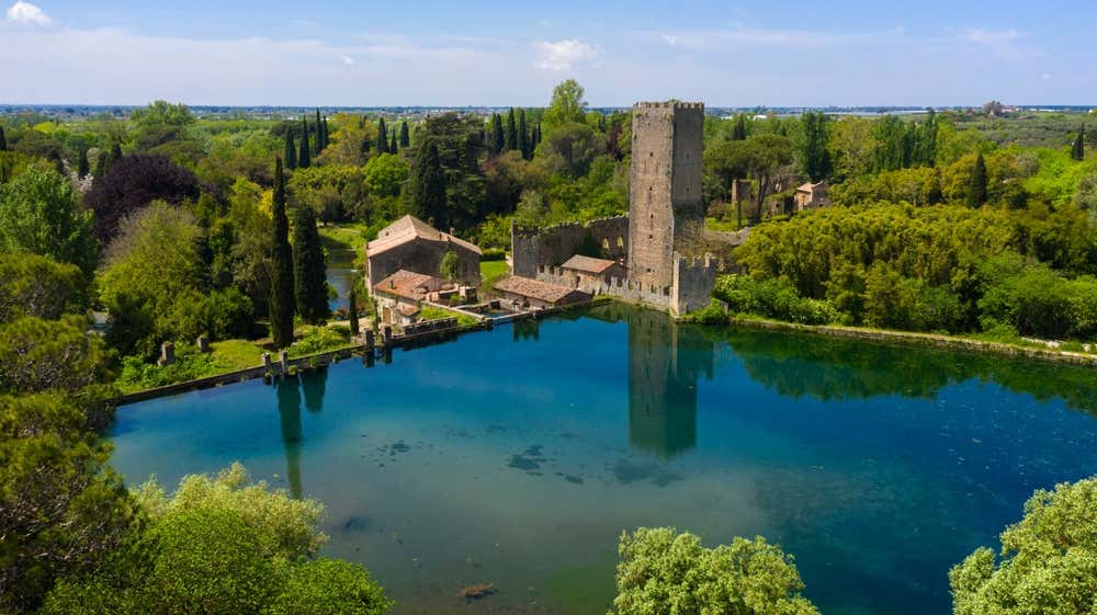 Vista aerea del Giardino di Ninfa, in provincia di Latina. Una torre medievale in pietra e i resti delle mura si riflettono in un laghetto dall'acqua limpida e turchese, circondato da cipressi, alberi in fiore e prati verdi sotto un cielo azzurro