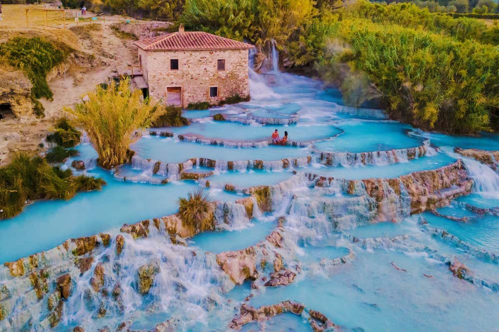 Vista aerea delle Cascate del Mulino di Saturnia, in Toscana: vasche naturali di acqua termale azzurra scendono a terrazze tra le rocce calcaree. In alto c'è l'antico mulino in pietra, con una piccola cascata accanto, e in una delle vasche si vede una coppia immersa nell'acqua. Tutto intorno cresce una vegetazione verde e dorata