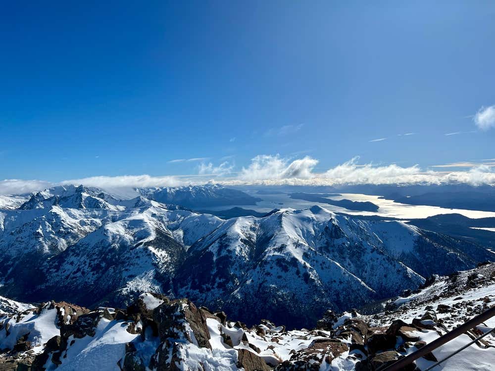 Vista panorâmica do Cerro Catedral coberto de neve, com o Lago Nahuel Huapi e as montanhas ao redor sob um céu com nuvens