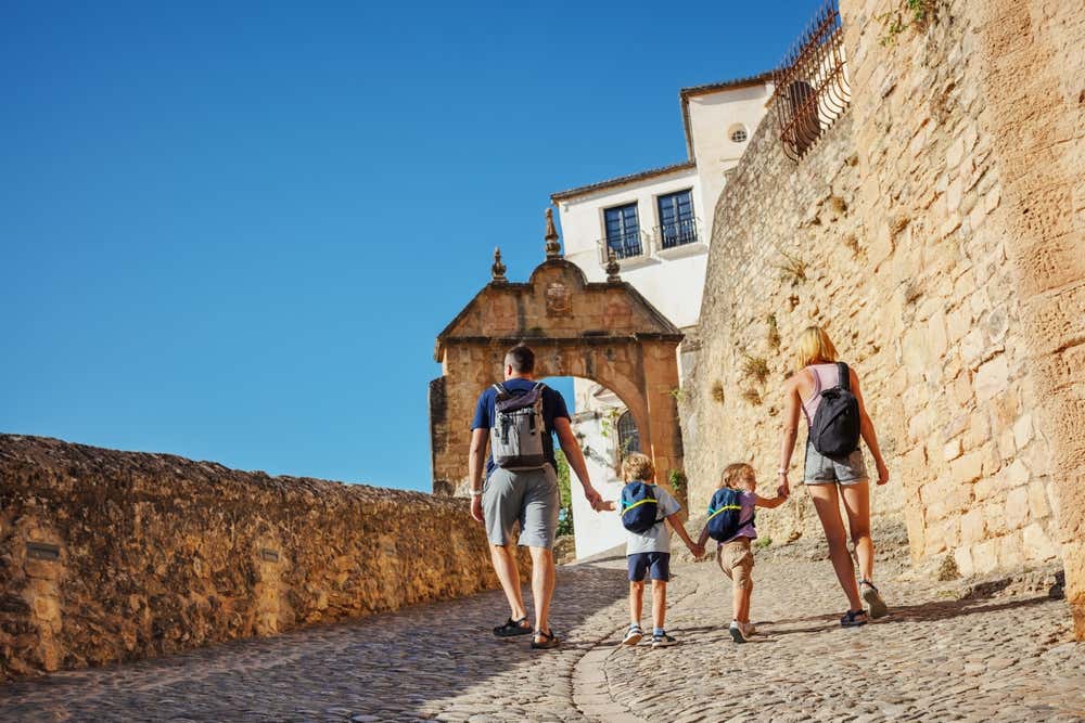 Una famiglia con due bambini e zaini in spalla cammina mano nella mano lungo una stradina in salita in ciottoli, diretta verso una porta ad arco in pietra a Ronda, un borgo storico spagnolo