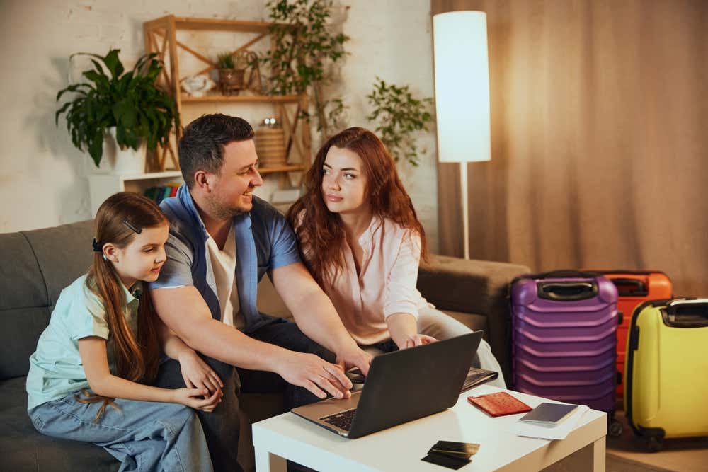 Una famiglia con una bambina, seduta sul divano di casa, pianifica una vacanza al computer portatile. Sul tavolino ci sono passaporti e documenti di viaggio, e accanto al divano sono pronte tre valigie colorate: viola, arancione e gialla
