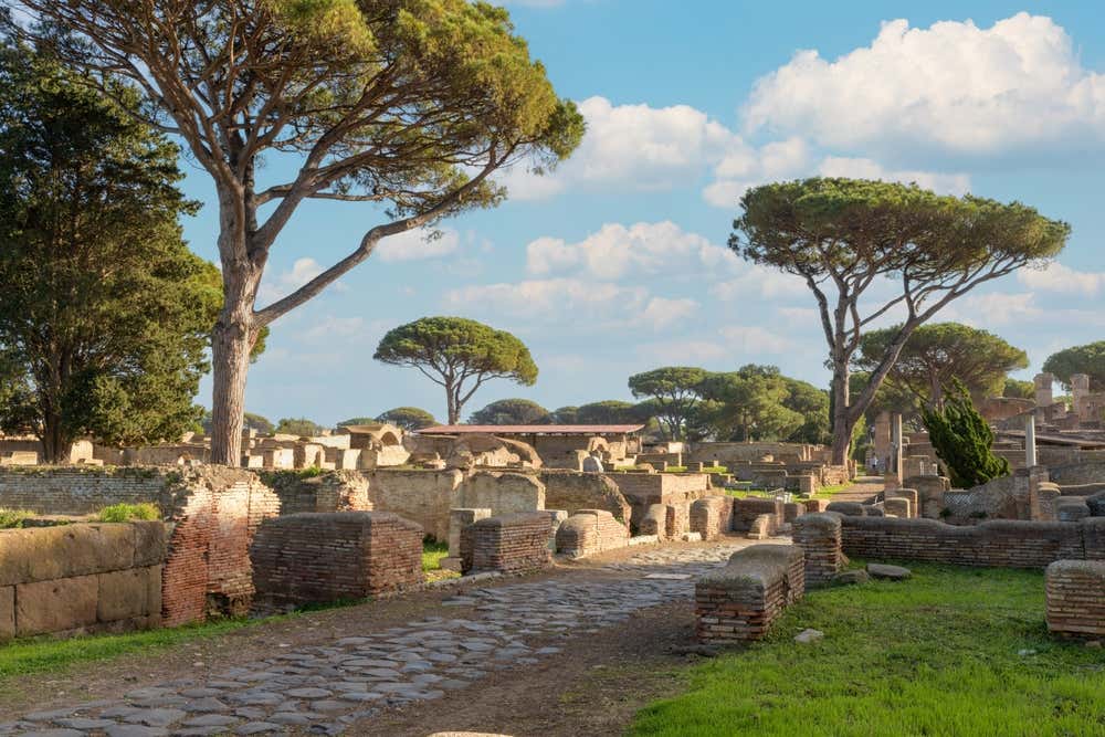 Gli scavi di Ostia Antica, vicino a Roma: un'antica strada romana lastricata attraversa i resti di edifici in mattoni. Tutto intorno ci sono pini marittimi e prati verdi, sotto un cielo azzurro con nuvole bianche