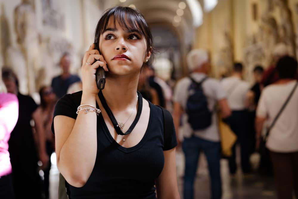 Una ragazza ascolta un'audioguida all'interno di un museo, in una galleria affollata di visitatori con sculture classiche e soffitti decorati sullo sfondo