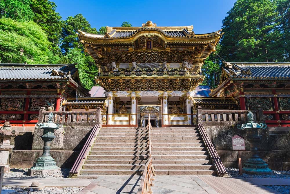 Lo Yomeimon, la porta monumentale del santuario Toshogu di Nikko, in cima a una scalinata in pietra. La porta è bianca e oro, ricoperta di intagli dipinti, con un doppio tetto dalle linee curve. Ai lati ci sono lanterne in bronzo ed edifici laccati di rosso, circondati da alti cedri sotto un cielo azzurro