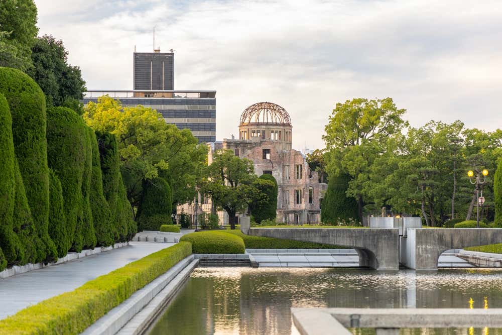 La Cupola della bomba atomica a Hiroshima, con la struttura metallica della cupola a vista e le mura in rovina, vista dal Parco del memoriale della pace. In primo piano ci sono una vasca d'acqua e siepi potate con cura, con alberi verdi e un edificio moderno sullo sfondo