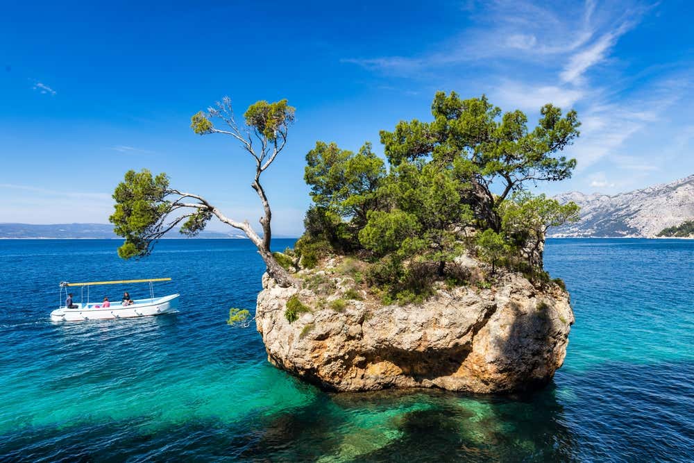 Lo scoglio di Brela (Kamen Brela), un grande faraglione roccioso ricoperto di pini marittimi che emerge dal mare cristallino della spiaggia di Punta Rata, con una barca a motore bianca nelle vicinanze